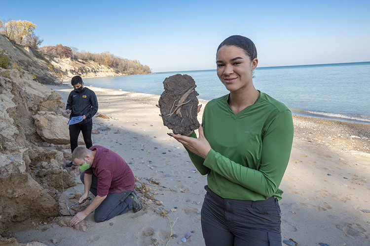 Photo of UWM student Mikayla Walker showing a fragment of peat with well-preserved plant and insect material unearthed at Sheridan Park in Cudahy. The fossil was found at the bottom of a bluff containing sedimentary strata deposited under a nearly mile-thick glacial at the end of the last ice age. Behind Walker are student Rene Chavez and geosciences lecturer Scott Schaefer. (UWM Photo/Troye Fox)