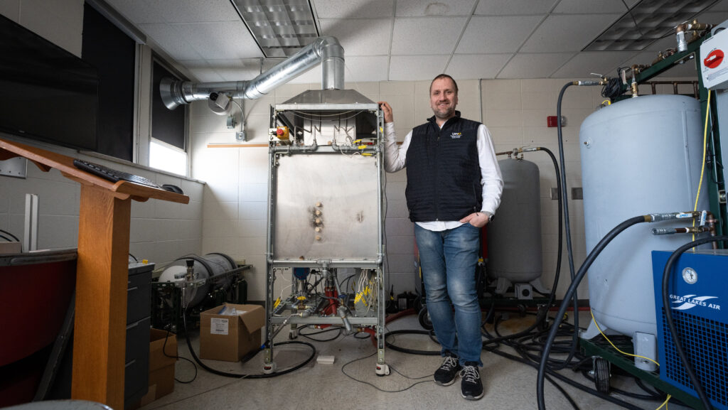 Photo of Olszewski posing next to the prototype of his flameless impingement oven, designed and built in the Teaching and Energy Research Industrial Lab on the Oshkosh campus. The oven recently was granted a U.S. patent. (Photo credit: UW Oshkosh)