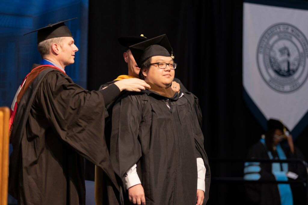 Photo of MFA design inspiring graduate Jackson Yang receiving his master's hood from Program Director Erik Evensen / UW-Stout