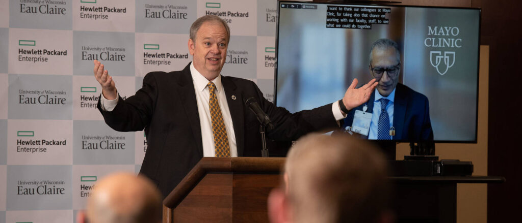 Photo of UW-Eau Claire Chancellor James Schmidt talking about a new public-private collaboration between Hewlett Packard Enterprise and the university at a Monday press conference. Pictured in the video screen is Mayo Clinic Health System Dr. Rajeev Chaudhry.
