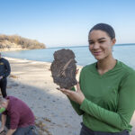 Photo of UWM student Mikayla Walker showing a fragment of peat with well-preserved plant and insect material unearthed at Sheridan Park in Cudahy. The fossil was found at the bottom of a bluff containing sedimentary strata deposited under a nearly mile-thick glacial at the end of the last ice age. Behind Walker are student Rene Chavez and geosciences lecturer Scott Schaefer. (UWM Photo/Troye Fox)