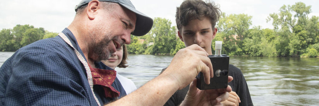 Photo of high school student Quienten Anger measuring nitrate value in the Chippewa River with Professor J. Brian Mahoney. Willow Anderson in the background.