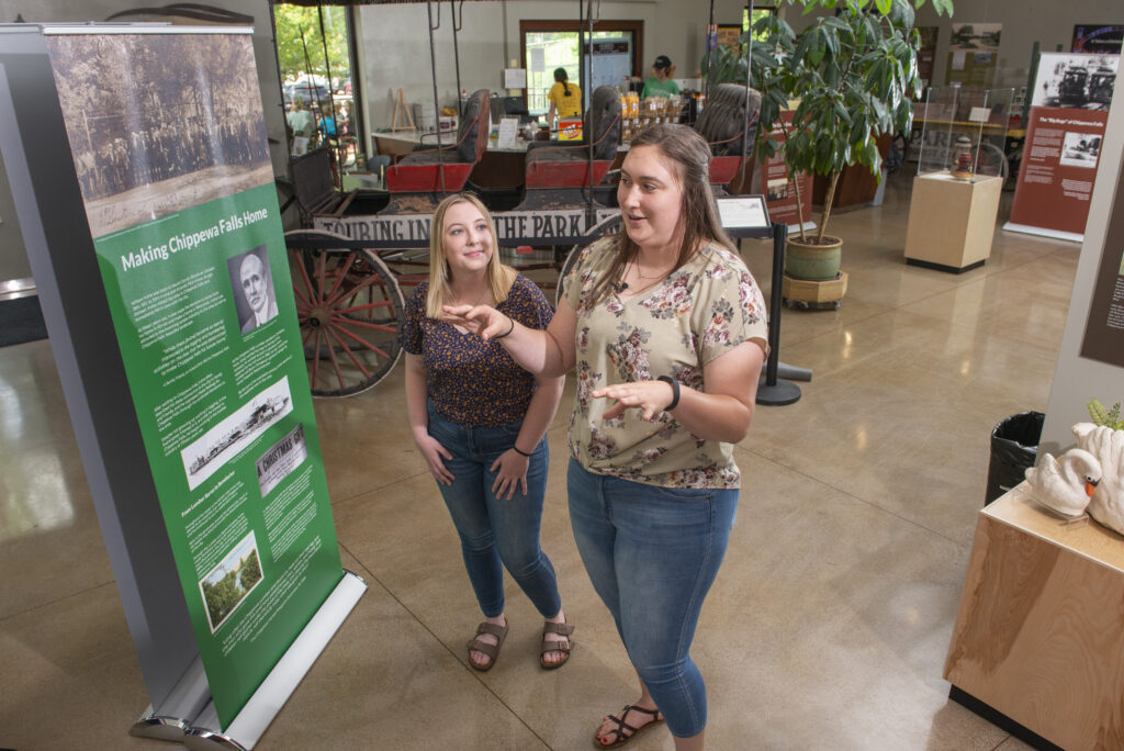 Photo of UW-Eau Claire public history graduate students Jordan Stish (left) and Alexi Linder discussing the exhibit they helped create in the Irvine Park Welcome Center in Chippewa Falls. (Photo by Shane Opatz)