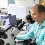 Photo of Ionel Popa, UWM professor of physics, demonstrating the magnetic tweezers built by his lab members to measure the mechanical forces that act on proteins as they fold and refold. Proteins are large molecules that carry out the body’s functions required for good health. In back is research assistant Sabita Sharma. (UWM Photo/Elora Hennessey)