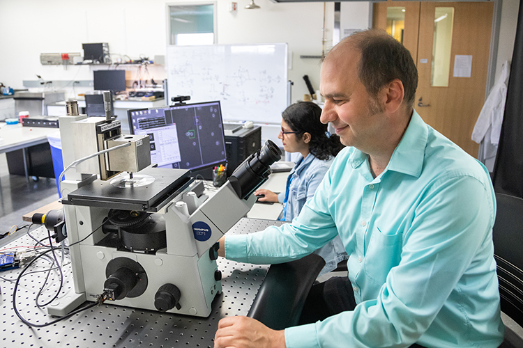 Photo of Ionel Popa, UWM professor of physics, demonstrating the magnetic tweezers built by his lab members to measure the mechanical forces that act on proteins as they fold and refold. Proteins are large molecules that carry out the body’s functions required for good health. In back is research assistant Sabita Sharma. (UWM Photo/Elora Hennessey)