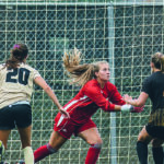 Photo of Elaina LaMacchia (in red) making a save during a UWM match. LaMacchia led the nation in goals-against average in 2021. (Milwaukee Athletics photo)