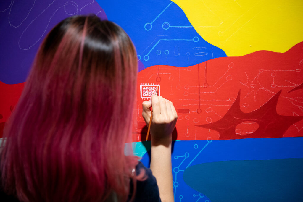 Photo of artist Sharon Tang painting a QR code on the mural, which depicts 12 real-life scientists as well as unnamed figures giving representation to minority voices not often heard in STEM. MICHAEL P. KING