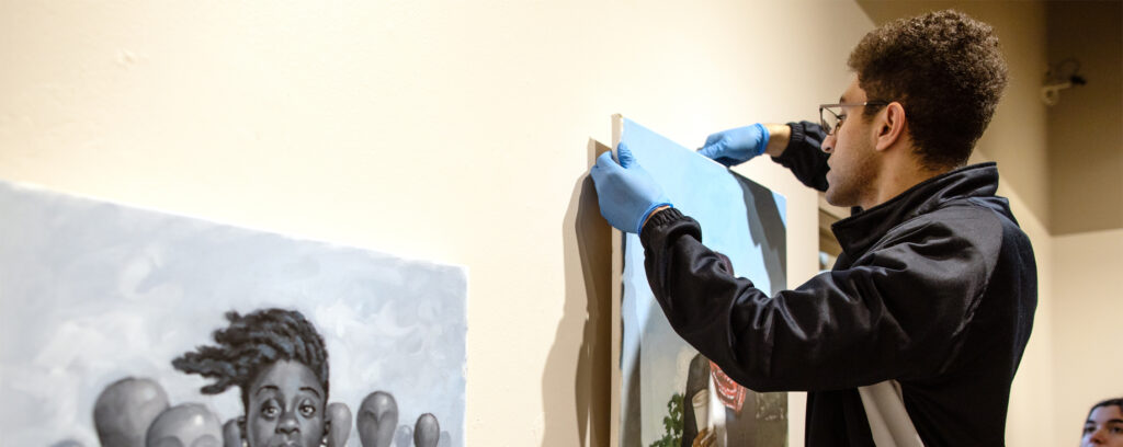 Photo of Brian Michael, a UW-Whitewater media arts and game development student from Sycamore, Illinois, hanging 12 works by painter Jerry Jordan at Roberta's Art Gallery on Jan. 25, 2023.