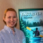 Photo of Megan Strom, graphic design major at UW-Stevens Point, posing with a travel poster she created for The Flow Project, which paired student artists and water professionals to create water-inspired art.