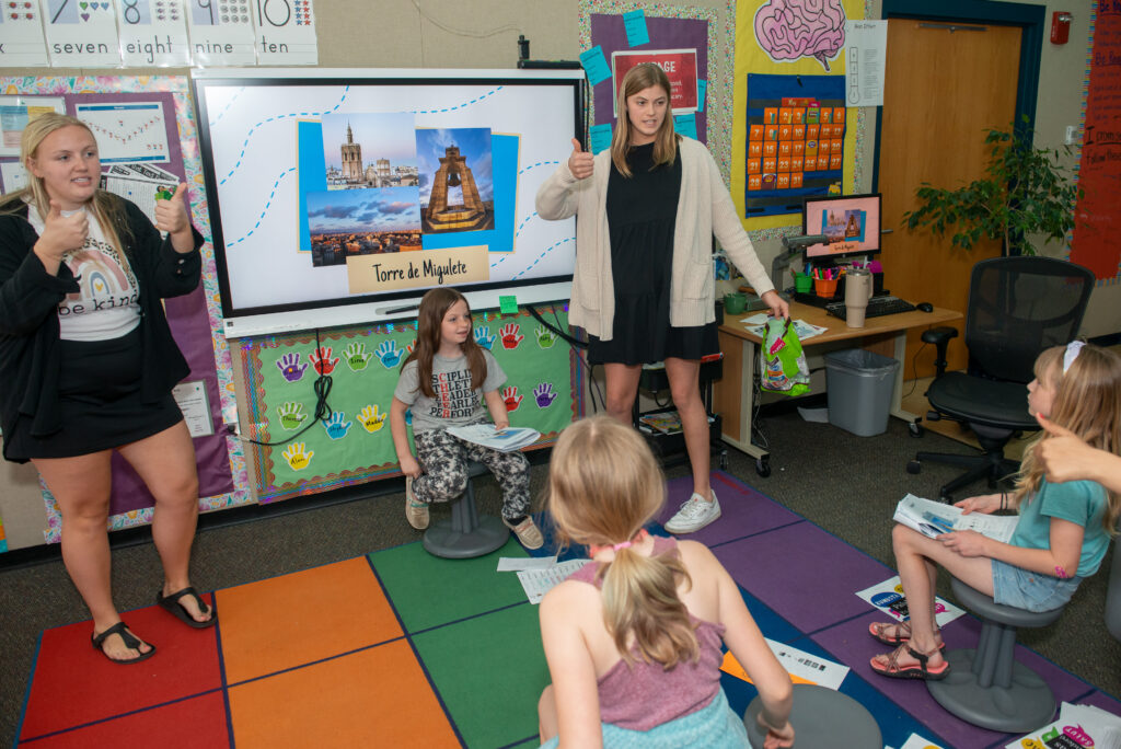 Photo of Halle Nicolet and Marli Felicijan, both senior Spanish education majors, shown here leading a class of young students through a lesson about spending a day in Valencia, Spain. At the end of the seven classes, the elementary school students presented the topic in Spanish to visiting parents.