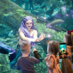 Photo of Pierce VanValkenburg as Mermaid Echo interacting with children attending an educational performance at Discovery World. (UWM Photo/Elora Hennessey)