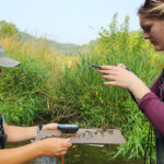 Photo of El Bartho (right) and Myah Eliason documenting curly leaf pondweed, an invasive aquatic species.