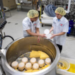 Photo of Brianna Crotteau, project coordinator at the Wuethrich Family/Grassland Dairy Center of Excellence at UW-River Falls, handing Sam Galgowski, a cheesemaker at the dairy plant, a plastic container that Galgowski filled with cheese during production on Tuesday. The cheddar cheese produced was the first produced at the dairy plant that will serve as a production site and a training location for students and dairy industry employees. UWRF/Pat Deninger photo.