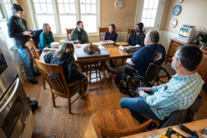 Photo of Farmers Jan Shepel (at near left) and Jim Koch (in wheelchair) flanking a 110-pound iron meteorite sitting on their family dining table at their home and Vienna EqHo Farm in the Town of Vienna, Wis. Included in the background, from left to right, are Shepel’s sister, Laurie Shepel; Carrie Eaton, curator of the UW Geology Museum; science writer Will Cushman; Noriko Kita, a distinguished scientist and meteorite expert from UW–Madison; and UW Geology Museum Director Rich Slaughter. At near right is Joe Zanter, a metallurgical engineer and Laurie Shepel’s husband. Photo: Jeff Miller