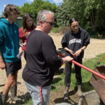 Photo of New Richmond High School agriscience teacher Rachel Sauvola shoveling corn from a gravity box as she instructs students, from left, Michael Mast, Jazzmira Traynor and Melany Rodriquez how to remove corn to be fed to animals at the school-operated farm that Sauvola began and oversees. Sauvola, a 1999 UW-River Falls graduate, was recently recognized for her teaching expertise by being named a 2025 Wisconsin Teacher of the Year. UWRF photo.