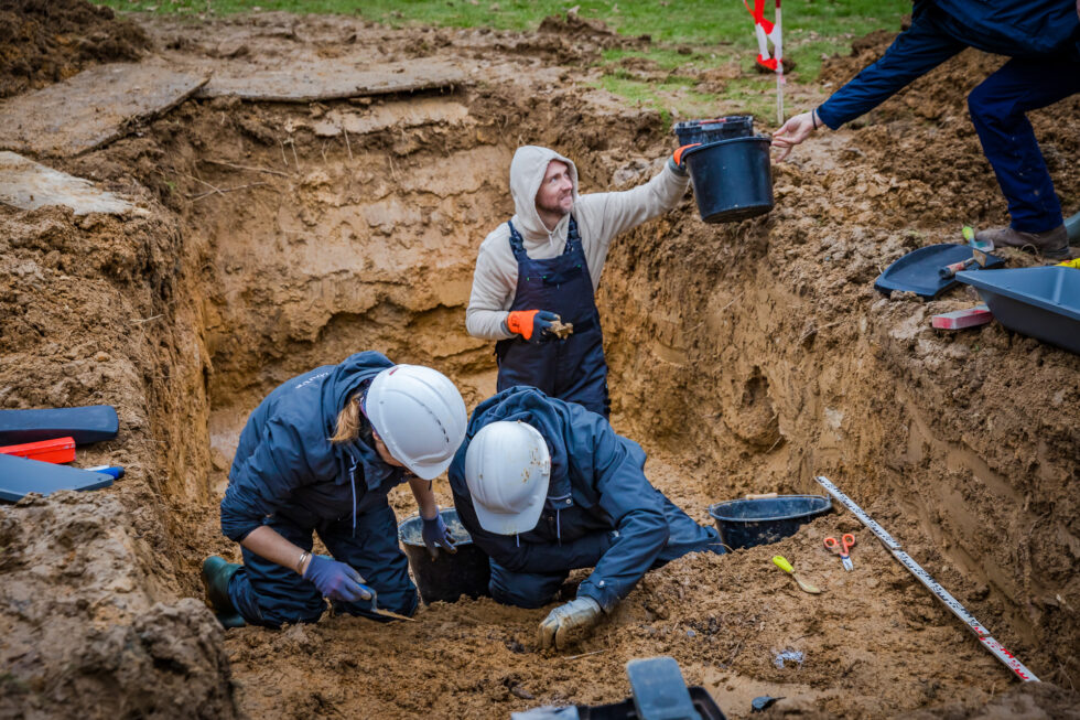 Photo of work at the site of a mass grave in Normandy, France, in December 2023. UW Oshkosh associate professor Jordan Karsten hands off a bucket containing bones to be submitted for DNA analysis. Photo courtesy Anibas Photography