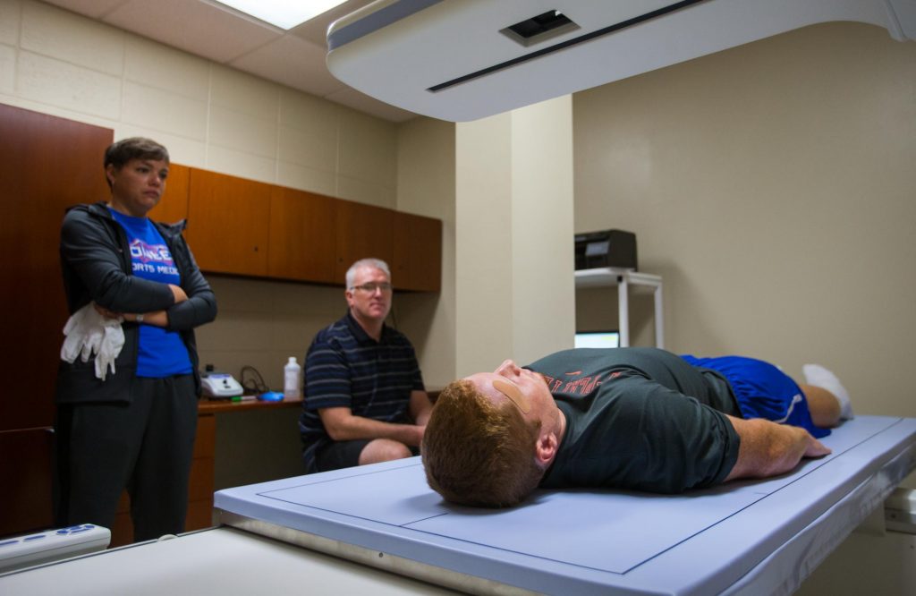 Scott Soja (sitting) performs a Dual Energy X-ray Absorptiometry (DXA) scan on a football player before a game to measure his muscle mass. Ryan Breckenridge (standing) is the team's head certified athletic trainer.