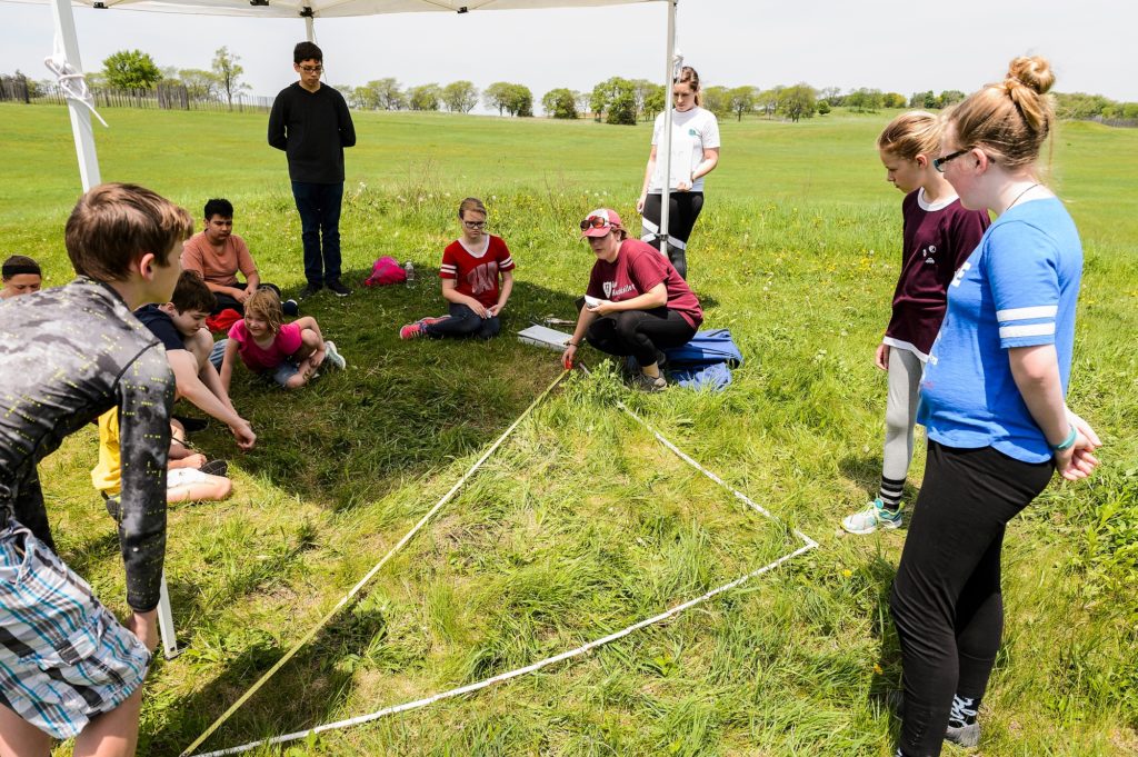 Photo of recent UW-Madison alumna Sarah Taylor, squatting at center, and Aleesha Kozar, standing at center, demonstrating a method for accurately measuring square grids of land as groups of sixth graders visiting from Fort Atkinson Middle School tour one of four educational stations temporarily set up at Aztalan State Park, a prehistoric Native American site located near Lake Mills, Wis., during spring on May 24, 2018. Funded by a Baldwin Wisconsin Idea Project Grant, the public outreach program is led by UW-Madison Anthropology Professor Sissel Schroeder, who has previously conducted several undergraduate archeological fields schools and research projects at the site. (Photo by Jeff Miller / UW-Madison)