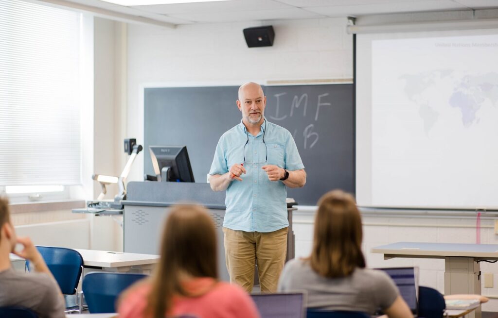 Male professor wearing a blue shirt and khakis stands at the front of a white-walled classroom speaking to a group of seated college students.