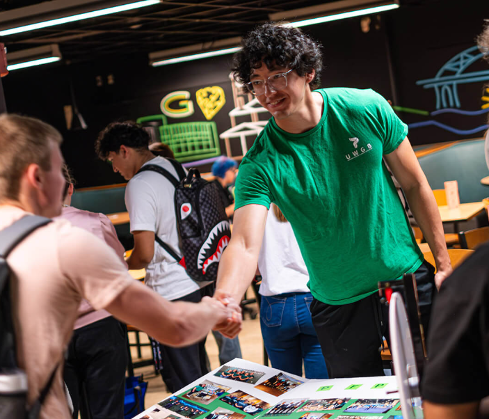 Male student wearing a green shirt and glasses shakes hands with another student.