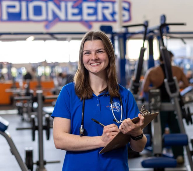 A female student holds a clipboard while wearing blue scrubs and a stethoscope.