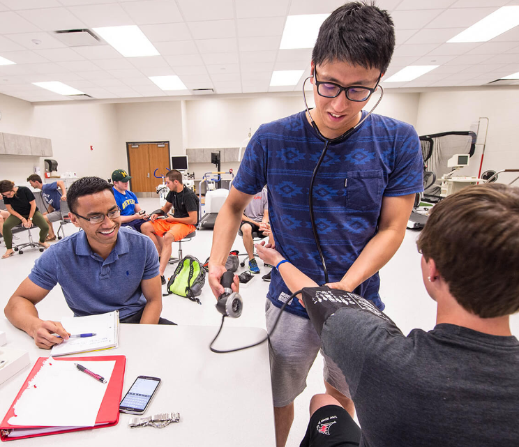 A male student wearing a blue shirt and glasses uses a stethoscope and blood pressure cuff to perform practice vitals check on a fellow student.