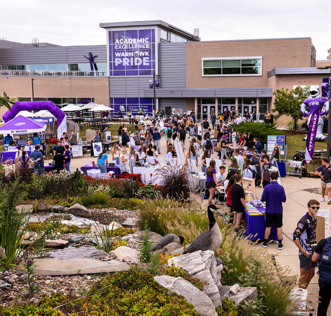 Outdoor view of a UWW academic building while a resource fair is hosted outside with students wearing campus gear and interacting with various tables.