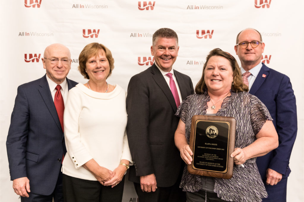 Photo of Elaina Koltz (holding plaque) receiving a Board of Regents 2019 Academic Staff Excellence Award; also pictured (from left) UW System President Ray Cross, Regent Janice Mueller, Regent Drew Petersen, and Regent Scott Beightol