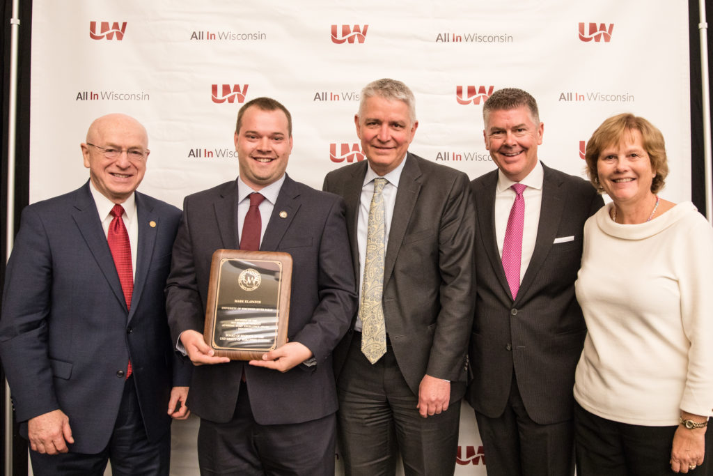 Photo of Mark Klapatch (holding plaque) receiving a Board of Regents 2019 Academic Staff Excellence Award; also pictured (from left) UW System President Ray Cross, Regent Bob Atwell, Regent Drew Petersen, and Regent Janice Mueller