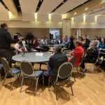 About 15 people seated at round tables with Ian Stroud of UW-River Falls standing and gesturing during a discussion session on Working with Students on Academic Recovery at the UW System Navigate and Advising Workshop March 9, 2023, at UW-Stevens Point