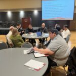 Several people seated at or around a round table during the Several people seated at 2 round tables with a projected PowerPoint slide in the background listing discussion topics including this one on Data Informed Practices at the UW System Navigate and Advising Workshop March 9, 2023, at UW-Stevens Point